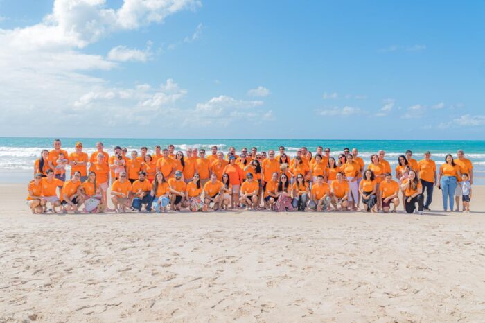 Grande grupo de pessoas vestindo camisetas laranjas reunido na areia da praia em Porto de Galinhas durante viagem de incentivo da BASF Top Consultores.